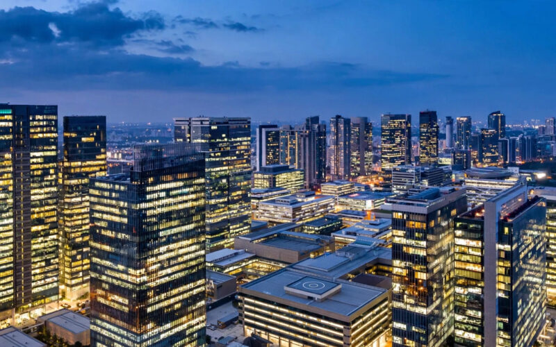 A skyline of modern glass office towers in Bengaluru at dusk with warmly lit windows against a deep blue sky.