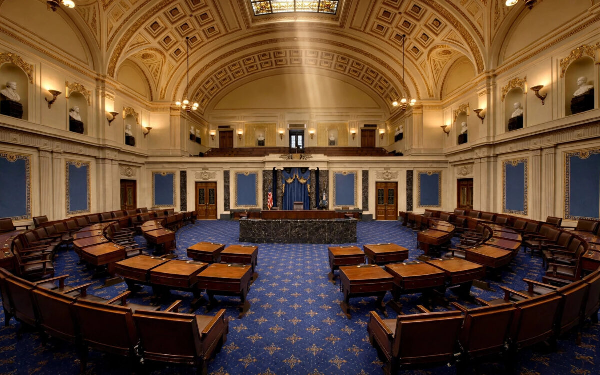 The empty US Senate chamber with wooden desks under a grand vaulted ceiling.
