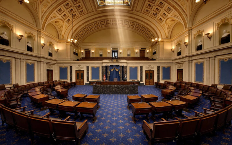 The empty US Senate chamber with wooden desks under a grand vaulted ceiling.