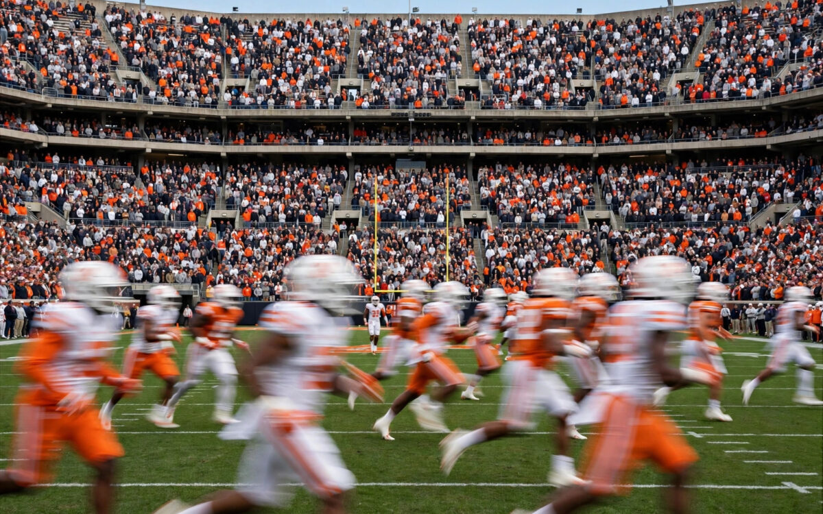 A packed college football stadium during a daytime game with players in motion on the field.
