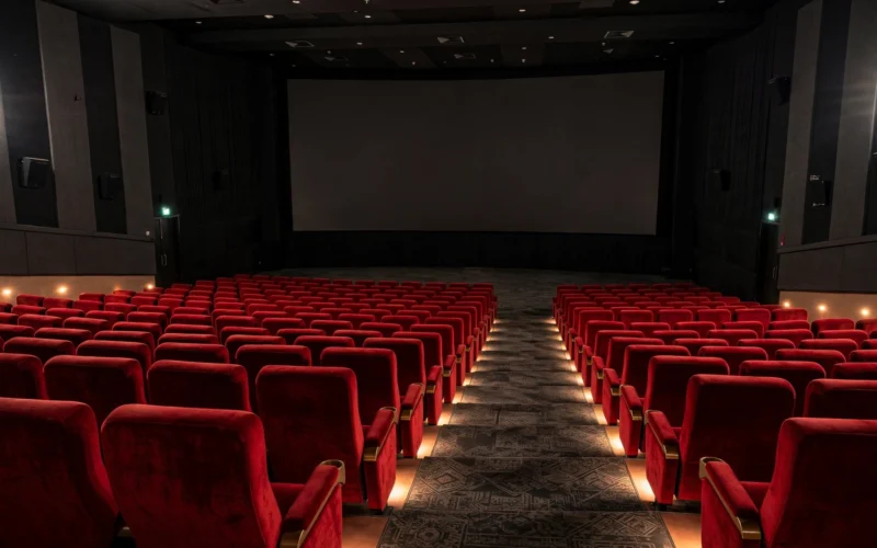 Interior of an Indian multiplex cinema hall with red seats facing a large dark blank screen.