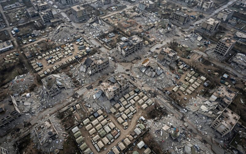 Aerial satellite-style view of destroyed city blocks with rubble and tent clusters