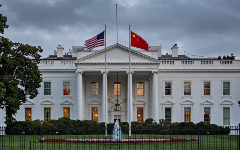 The White House stands at dusk with American and Chinese flags flying side by side under an overcast sky.